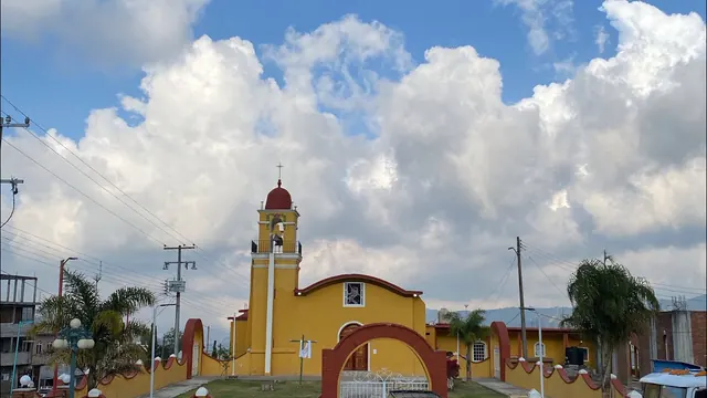Iglesia de fachada amarilla con campanario, bajo un cielo nublado.